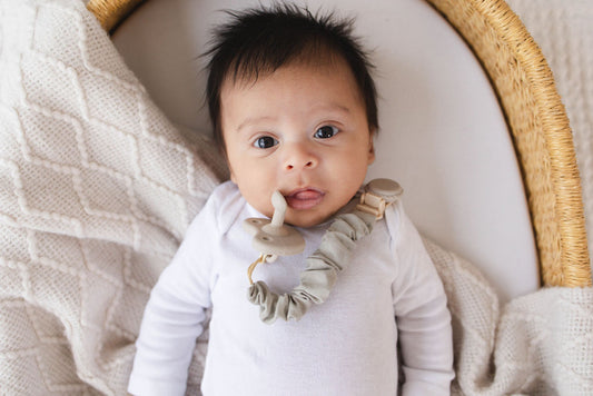 Newborn lying in a bassinet wearing a Mila & Lulu sage silicone pacifier and metal-free clip