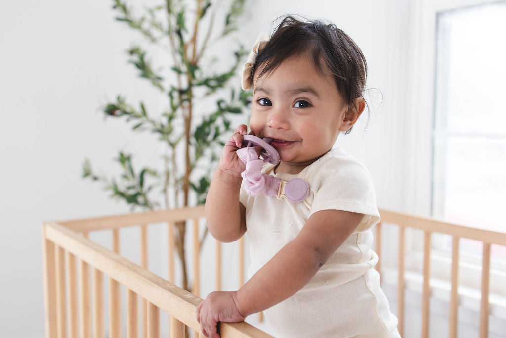 Toddler smiling in crib holding a Mila & Lulu mauve silicone pacifier with metal-free clip
