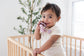 Toddler smiling in crib holding a Mila & Lulu mauve silicone pacifier with metal-free clip